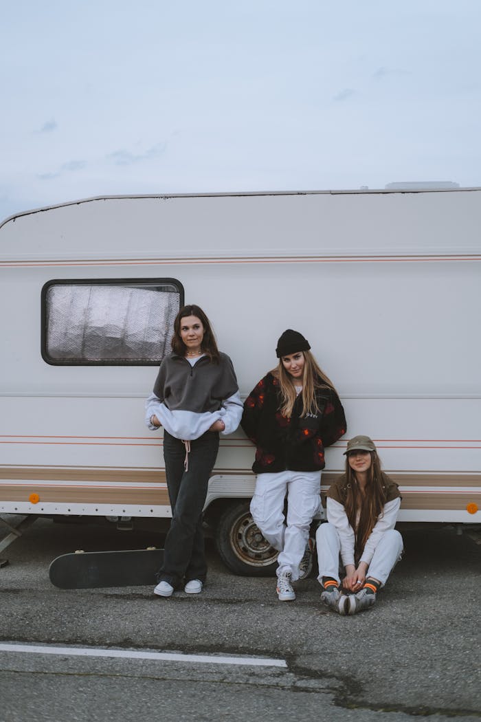 Three young women in casual streetwear stand and sit by a caravan, embodying street culture.